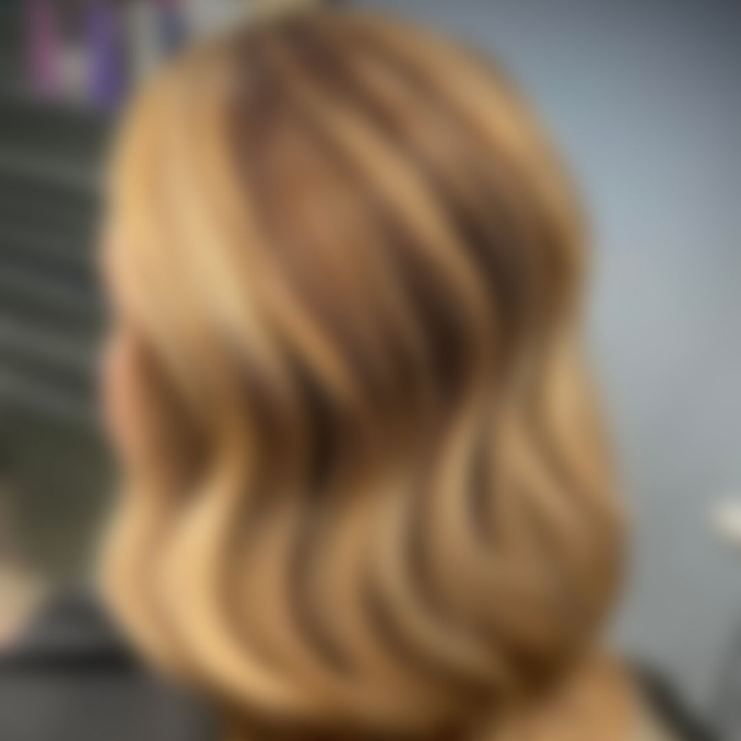 A blurred image showing the back of a person’s head with shoulder-length, wavy, blonde hair. The background appears to be indoors, possibly in a salon.